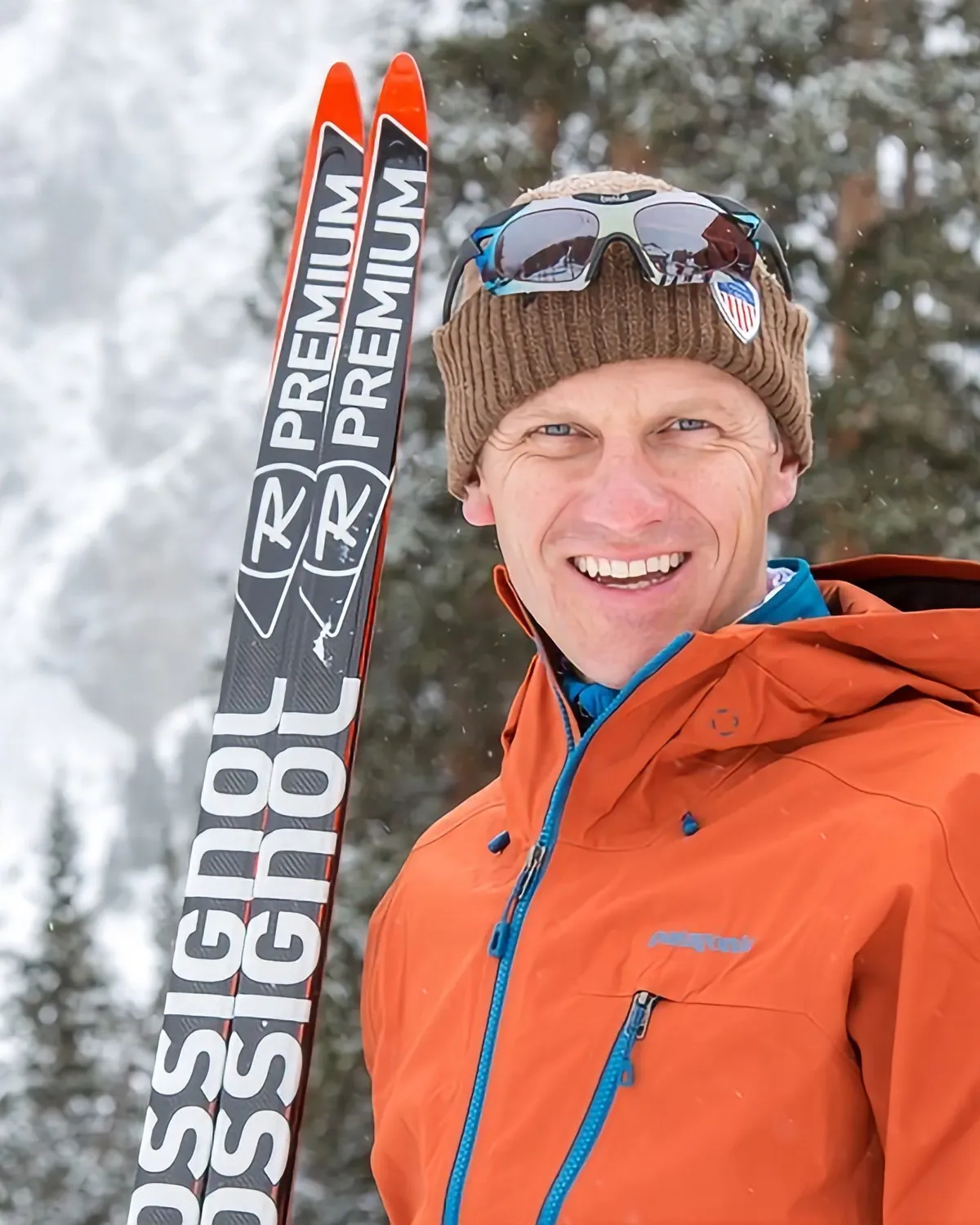 Coach Greg in an orange shell jacket and knit hat, smiling beside a pair of upright Rossignol skis with snowy pines behind him