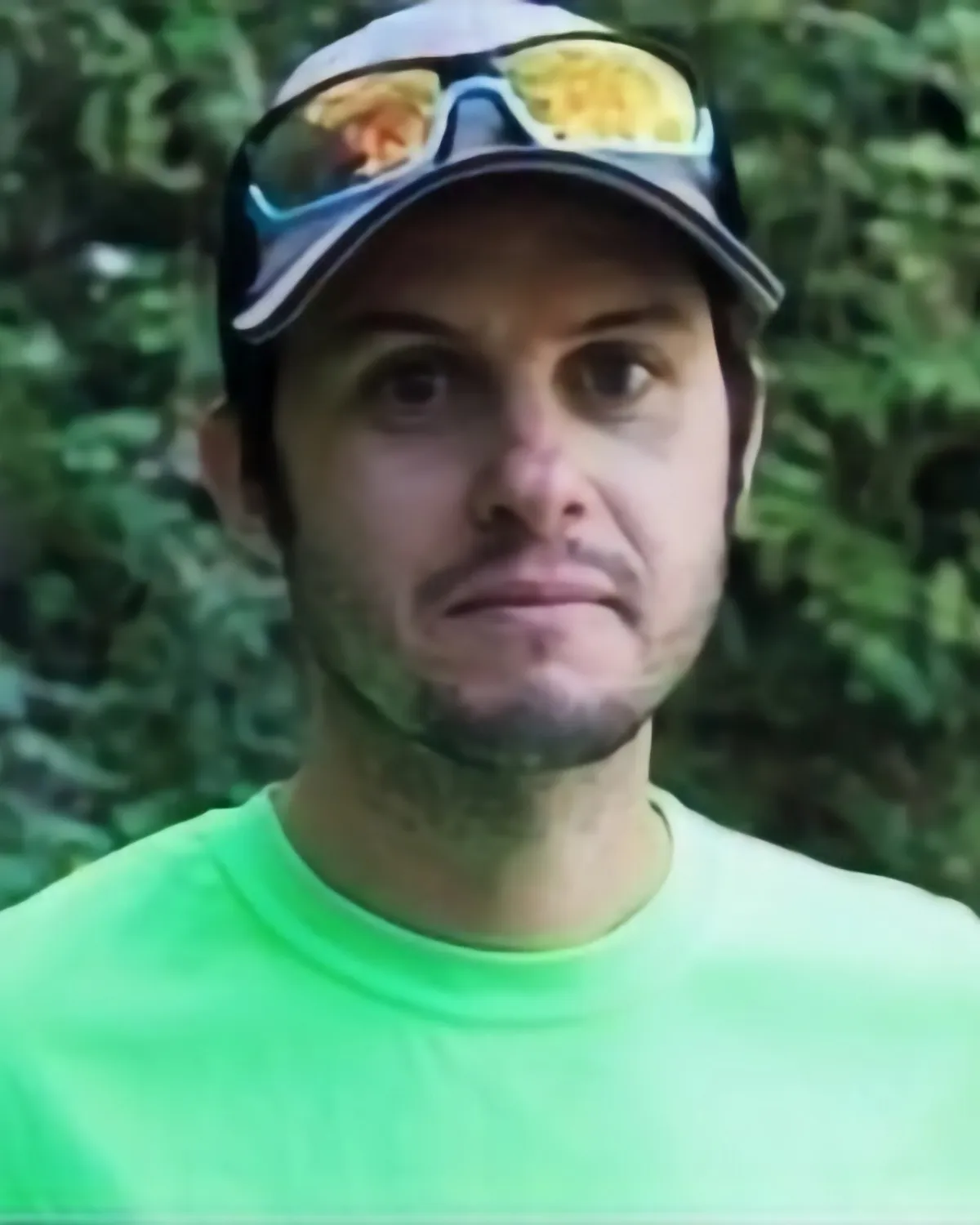 Coach KJ in a baseball cap with sunglasses perched on the brim, smiling in front of a leafy green backdrop
