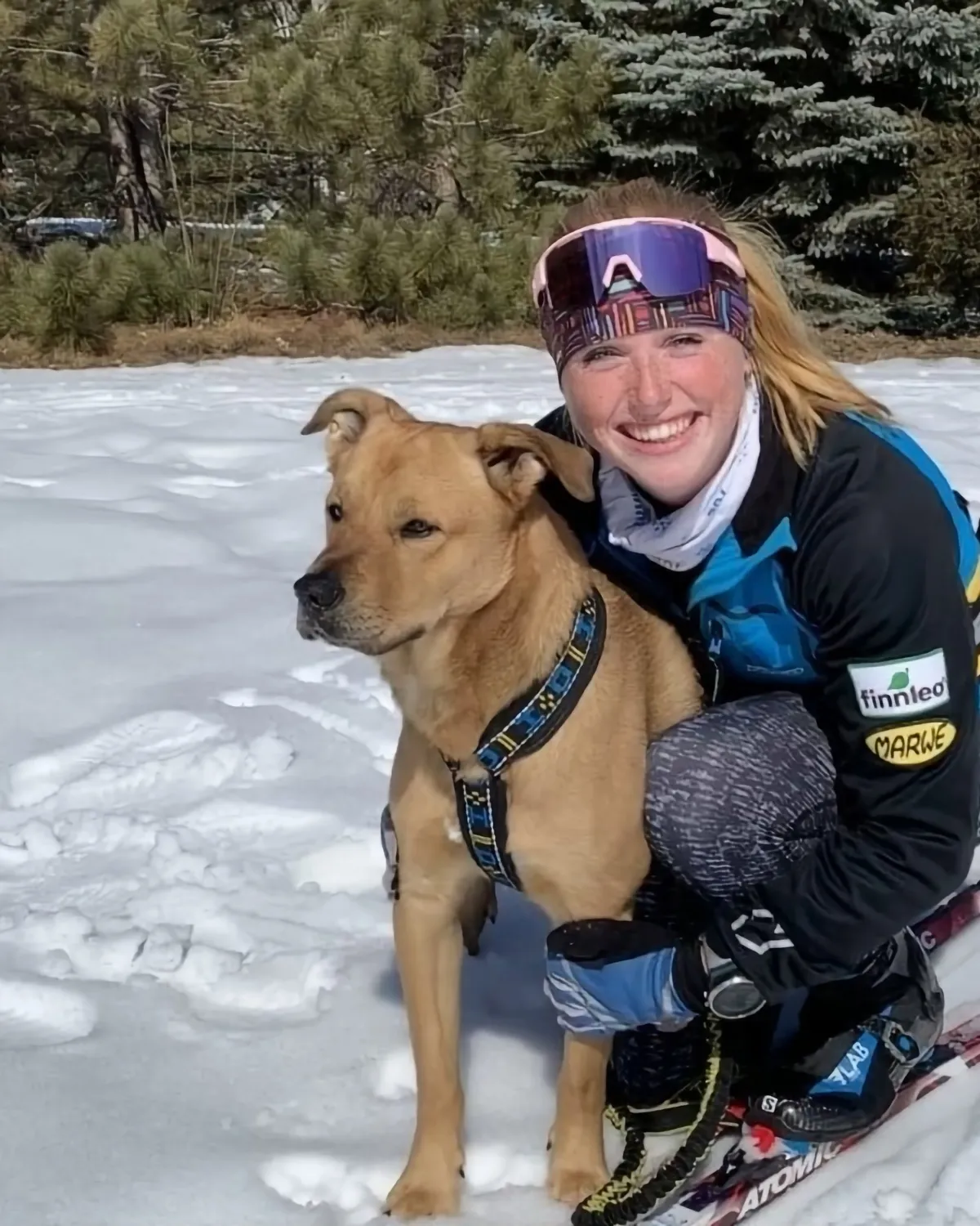 Coach Rebecca in a race suit and pink-framed ski goggles, kneeling on snow with her arm around a tan dog wearing a harness