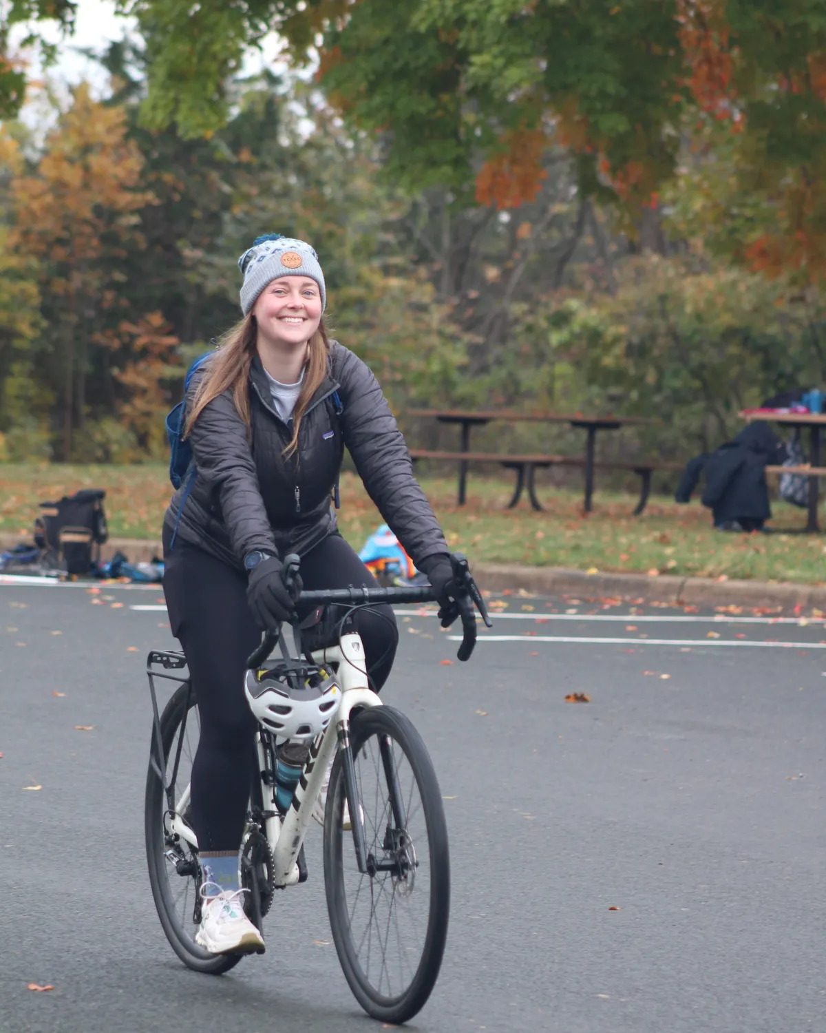 A smiling rider in a beanie on the bike leg, fall color behind her