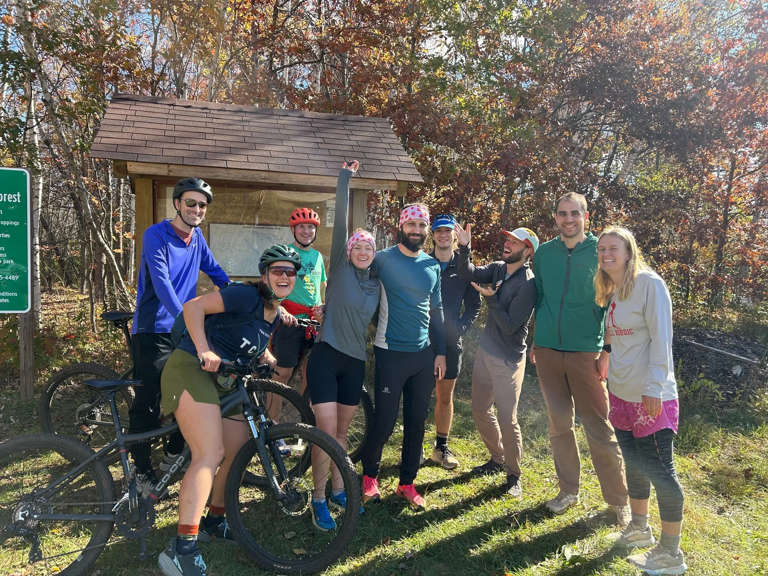 A grinning group of members gathered with a mountain bike at a wooded trailhead in fall color