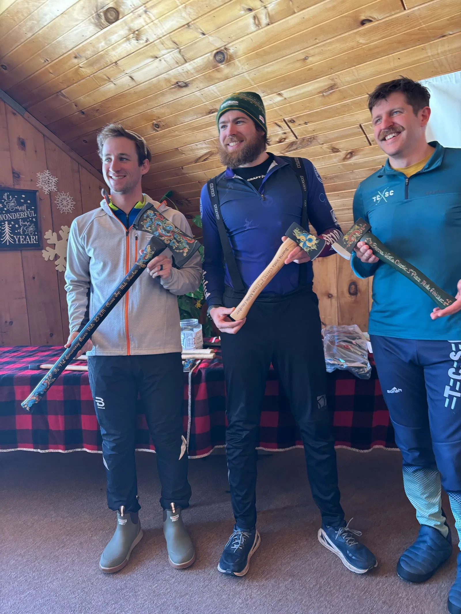 Three racers in a wood-paneled cabin holding painted axe trophies from the Finlandia