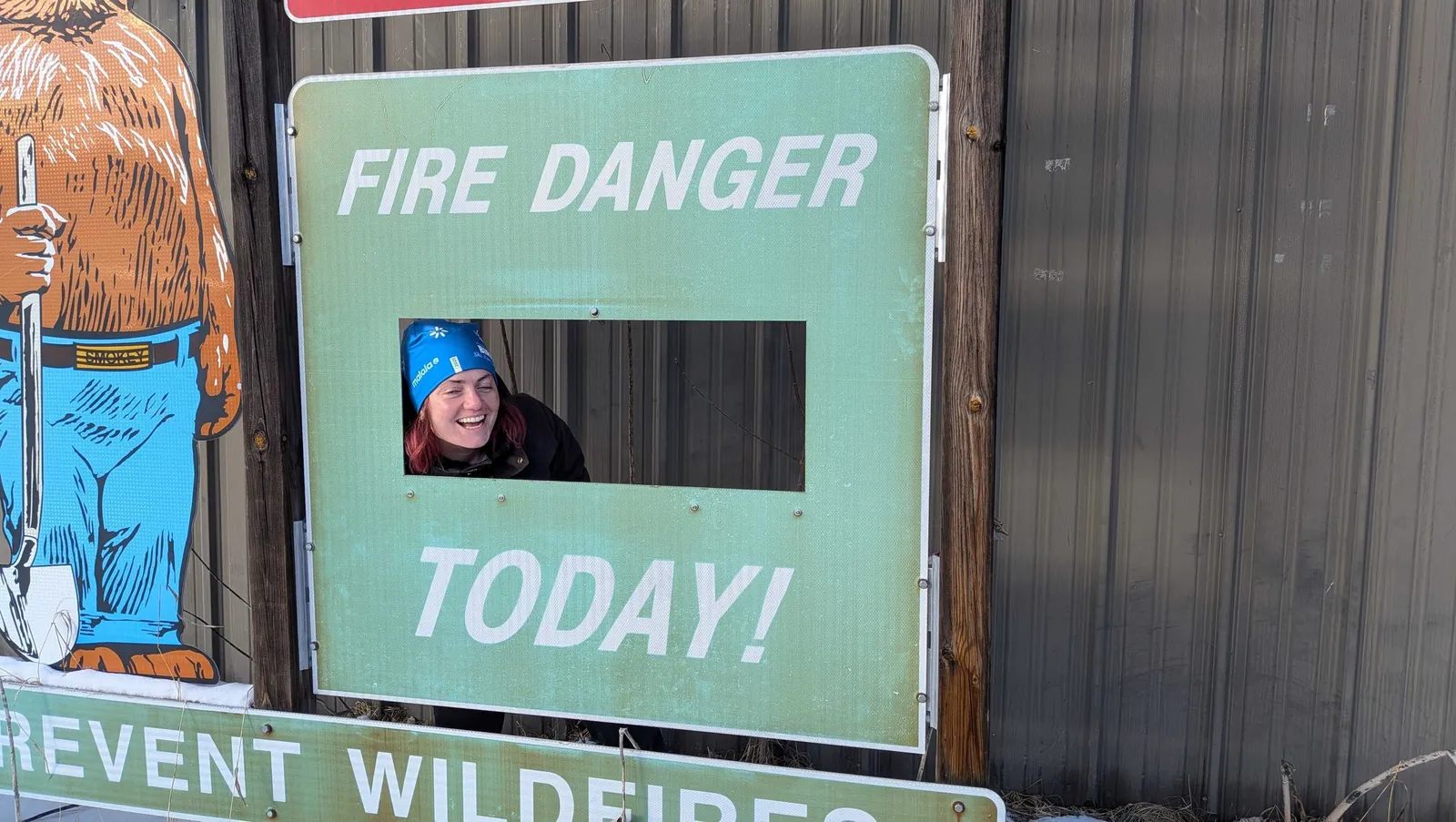 A member in a ski beanie grinning through the cutout of a Smokey Bear fire danger sign