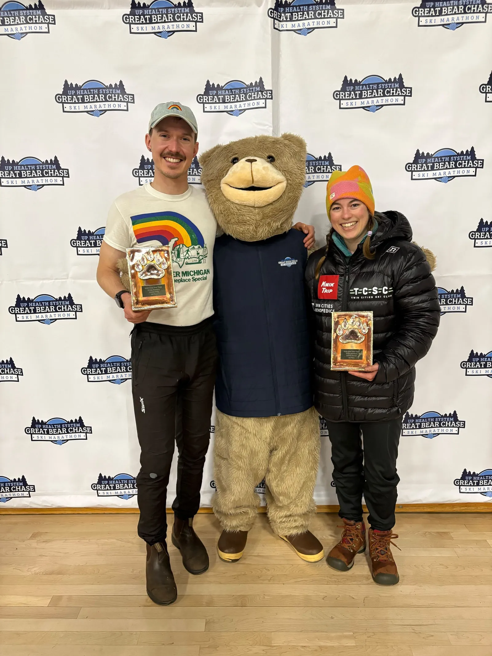 Two members holding wooden podium plaques on either side of the Great Bear Chase bear mascot
