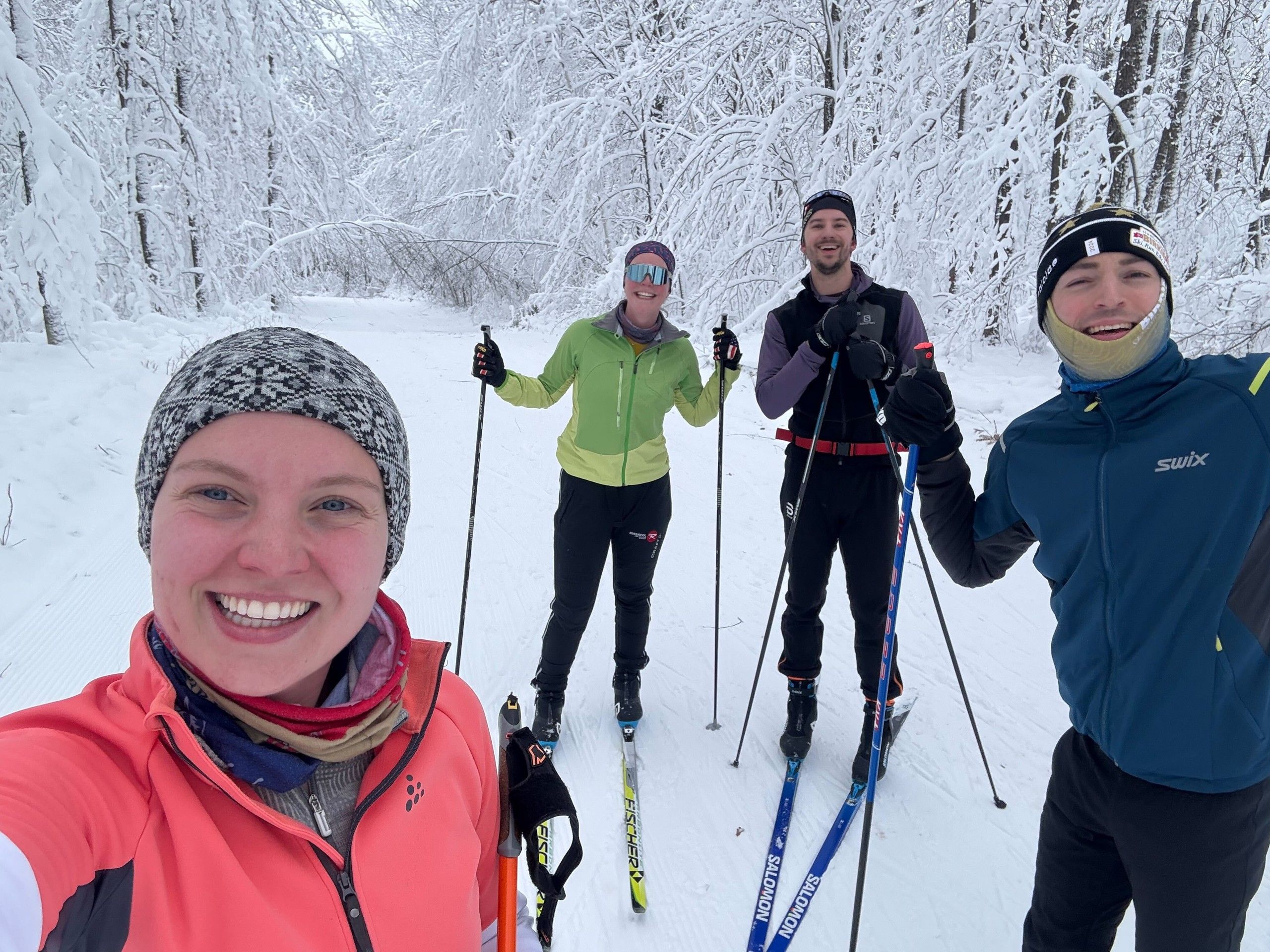 Four TCSC skiers grinning on a groomed trail through a forest completely coated in fresh snow, skis and poles in hand