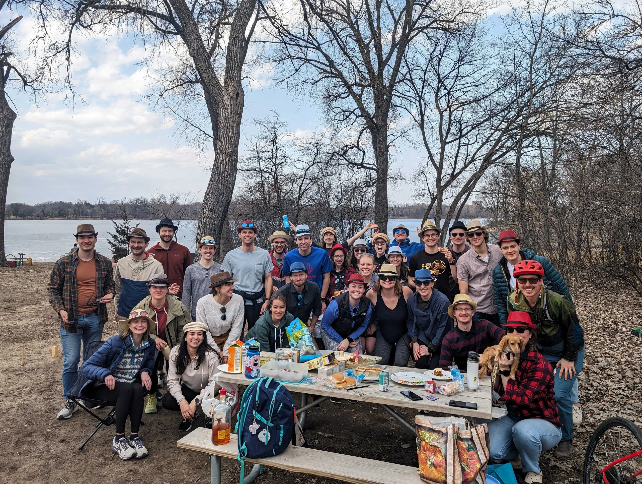 A lakeside picnic crowd in novelty hats around a packed picnic table