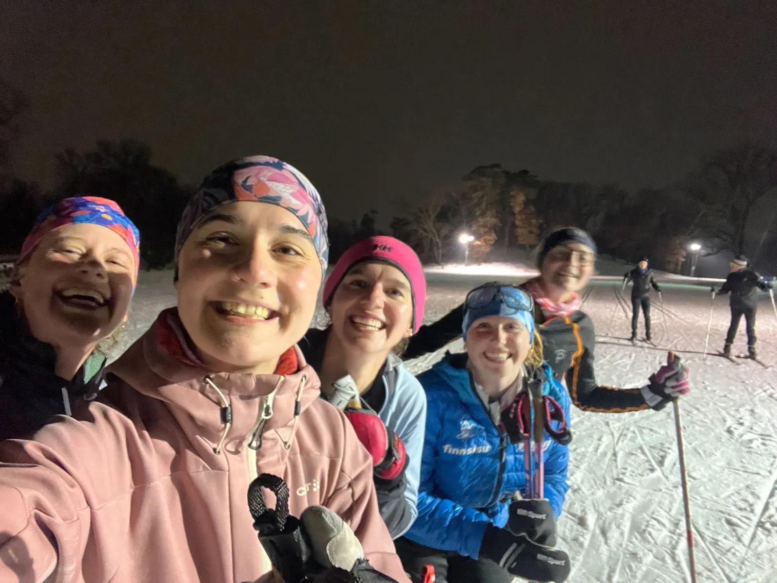 Five smiling skiers crowd into a selfie at a night practice, trail lights and classic tracks behind them