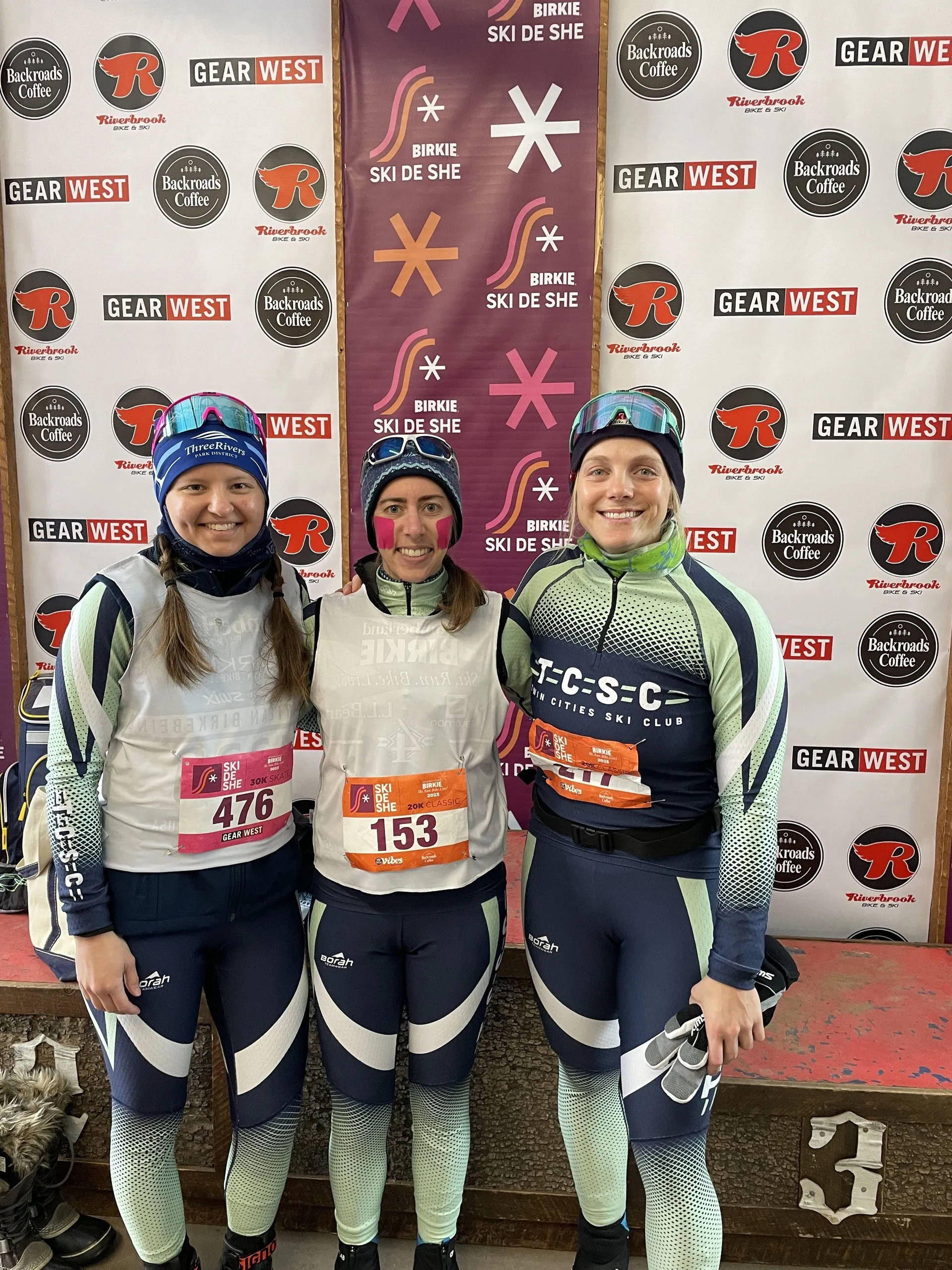Three members in TCSC jerseys and race bibs in front of the Ski de She sponsor wall