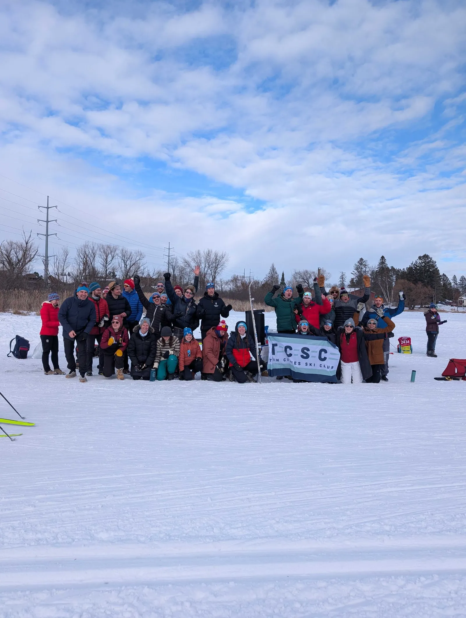 A few dozen members in winter layers gathered behind the TCSC flag on snow at the Birkie, arms raised