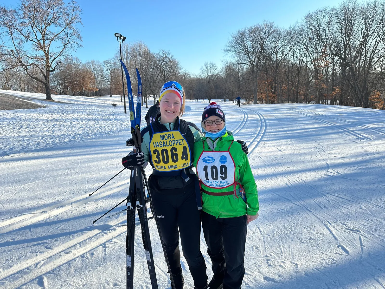 Two skiers wearing Mora Vasaloppet bibs side by side on a sunlit trail, skis in hand